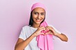 © Krakenimages.com - Young brunette woman wearing breast cancer support pink scarf smiling in love showing heart symbol and shape with hands. romantic concept.