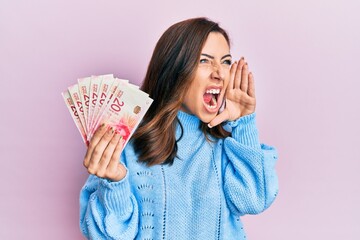 Canvas Print - Young brunette woman holding 20 israel shekels banknotes shouting and screaming loud to side with hand on mouth. communication concept.