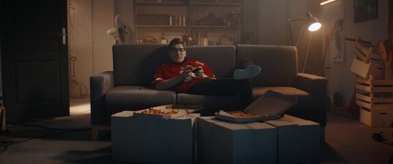 POV Portrait of Caucasian teenager playing video game inside home garage, enjoying pizza. Shot with 2x anamorphic lens