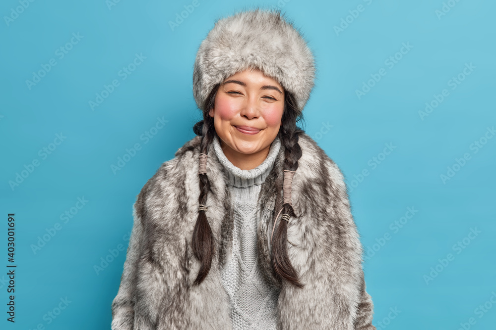 Winter clothes. Pleased Inuit woman with two pigtails smiles gently wears fur hat knitted warm sweater and coat poses against blue background. Happy nenets girl indoor. Aboriginal from north - obrazy, fototapety, plakaty 