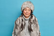 Winter clothes. Pleased Inuit woman with two pigtails smiles gently wears fur hat knitted warm sweater and coat poses against blue background. Happy nenets girl indoor. Aboriginal from north