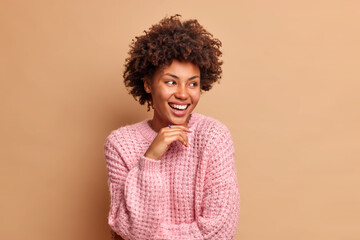 Wall Mural - Portrait of curly haired woman keeps hand under chin and looks away gladfully wears knitted jumper has carefree expression poses against brown background. Happy emotions and feelings concept
