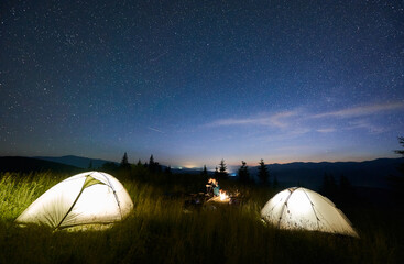 magnificent view of night starry sky over grassy valley with hikers and illuminated camp tents. man 