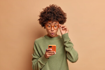 Photo of speechless impressed dark skinned curly beautiful woman stares at smartphone stands with bugged eyes keeps hand on rim of spectacles wears casual jumper has shocked expression reads news.