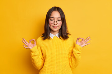 Wall Mural - Studio shot of of calm young Asian woman shows zen or okay sign meditates in good mood stands with eyes closed wears casual sweater and spectacles isolated over yellow background feels relieved