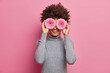 Photo of curly haired young ethnic woman covers eyes with pink gerberas going to make bouquet from flowers dressed in casual grey turtleneck isolated over rosy background. Professional florist