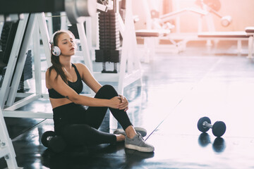 Picture of attractive fitness woman sitting on the floor at gym while using phone and listening music with headphones