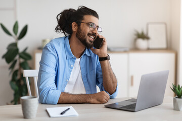 Young Arab Man Talking On Cellphone, Working On Laptop In Home Office