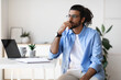 Thoughtful young arab male freelancer sitting at desk at home, looking aside