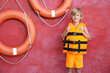 © New Africa - Little boy wearing orange life vest near red wall with safety rings