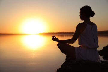 Wall Mural - Woman practicing yoga near river on sunset. Healing concept