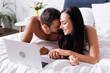 © LIGHTFIELD STUDIOS - happy couple laughing while holding hands near laptop on blurred background in bedroom