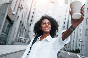 Wall Mural - With cup of drink. African american woman in good clothes is outdoors in the city at daytime