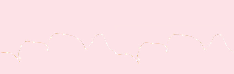 Partially defocused string of warm lights, garland on tender pink background, festive decoration or party wide banner. Selective focus