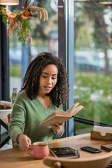 Poster - african american woman holding book and reaching mug with coffee in cafe