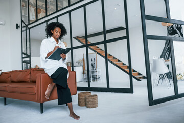 Wall Mural - Holds notepad. Young african american woman with curly hair indoors at home