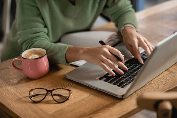 Wall Mural - cropped view of african american woman holding pen while typing on laptop keyboard near cup of coffee