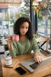© LIGHTFIELD STUDIOS - african american woman holding cup of coffee while using laptop in cafe