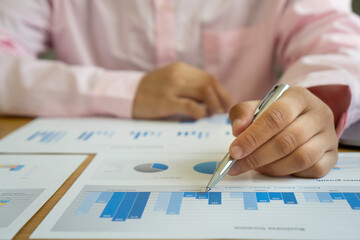 Asian finance staff is sitting at the table. The hand is holding a pen pointing to a graph document showing the company's growing profits, financial business ideas. Corporate profit growth