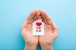 © fotoduets - White paper house with bright red heart in young adult woman palms on light blue table background. Pastel color. Closeup. Point of view shot. Top down view.