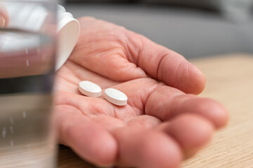 White pills of painkiller or antibiotic for treatment on senior woman hand palm, glass with water, medicines and vitamin supplements concept,close-up view