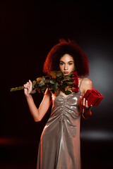 Wall Mural - curly african american woman in dress holding gift box and roses on black