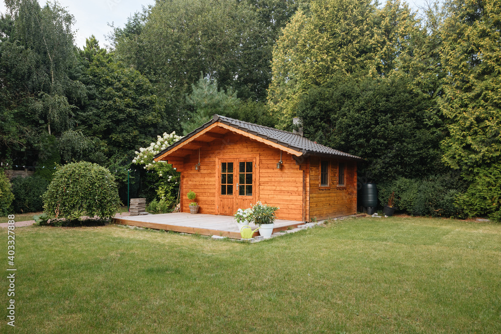 Orange wooden hut in the garden with many tall trees. Garden shed with lawn in front of him - obrazy, fototapety, plakaty 