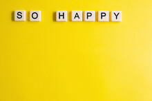 the words so happy written as a flat lay in wood scrabble tiles on a plain yellow background