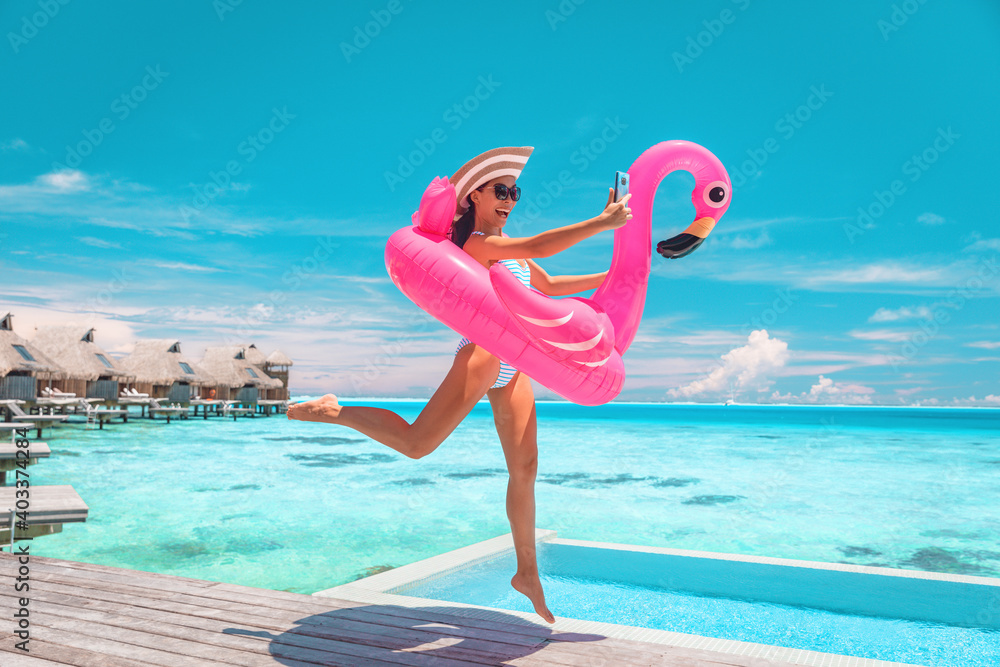 Happy fun luxury hotel vacation woman jumping of joy taking selfie with pink inflatable swimming pool mattress at overwater bungalow resort. - obrazy, fototapety, plakaty 