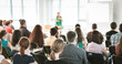 © kasto - Business and entrepreneurship symposium. Female speaker giving a talk at business meeting. Audience in conference hall. Rear view of unrecognized participant in audience.