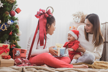 Wall Mural - parent and two little children having fun and playing together near christmas tree indoors. merry christmas and happy holidays. cheerful mom and her cute daughters girls exchanging gifts.