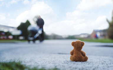 Wall Mural - Rear view Lonely Teddy bear doll sitting alone on hailstones with blurry father pushing stroller walking on footpath.lost brown bear toy looking out on the road, International missing children's day