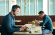 © Mint Images - Two men seated in a cafe, working remotely  using a  laptop and smart phone