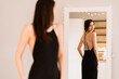 Woman in a long black dress with bare back looking at her reflection in the mirror while getting ready for an event, party, date
