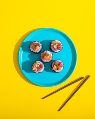 Wall Mural - Abstract sushi roll on blue plate on yellow background with chopsticks