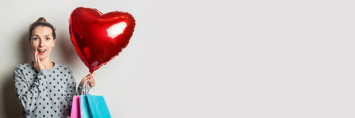 Surprised Young woman in a sweater holds packages for shopping and a heart air balloon on a light background. Valentine's Day concept. Banner