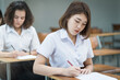 © EduLife Photos - Selective focus of the teenage college students sit on lecture chairs do final examination and write on examination paper answer sheets in the classroom. University students in uniform in classroom.