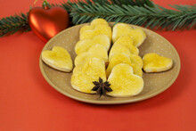 Many Pieces Of Heart-shaped Curd Cookies Covered With Yellow Jam Lie With An Aniseed Star In A Brown Plate On A Red Background On The Side Next To A Tree Branch And A Red Christmas Toy