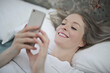 © olly - young woman lying on the bed writes with smartphone
