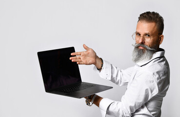 Stylish grey-haired bearded hipster man in eyewear pointing to black blank laptop screen looking at camera. Studio shot closeup portrait isolated on white background. Recommend, presentation