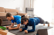 Couple doing abdominal planks in living room dressed in sportswear, brightly lit room and tablet beside