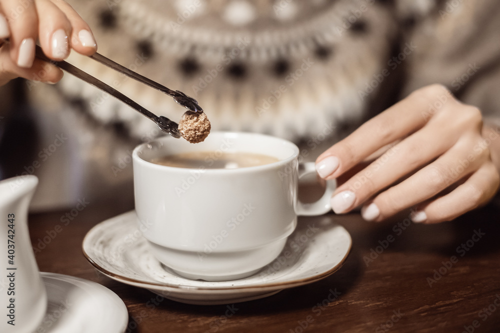Woman puts a piece of brown cane sugar in a mug of coffee. The concept of healthy eating and excess of glucose and fast carbohydrates - obrazy, fototapety, plakaty 