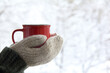 a red mug in the hands of those dressed in knitted mittens against the backdrop of a blurred snow landscape. a warming drink for a winter morning