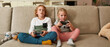 © Svitlana - Two siblings, little boy and girl looking focused, playing video games using joystick or controller while sitting together on a couch at home