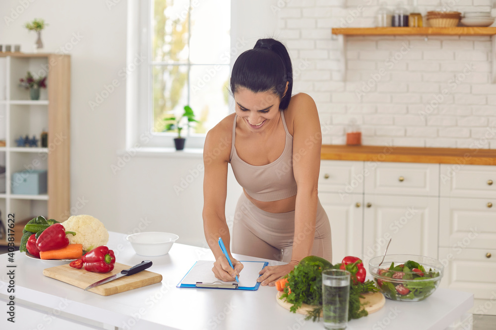 Cheerful pretty slim female athete in sportswear standing in kitchen in sportswear and writing down healthy recipe or daily ration diet at home. Active healthy lifestyle, clean eating concept - obrazy, fototapety, plakaty 