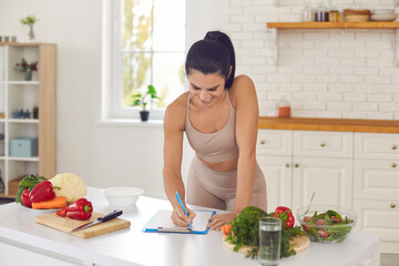 cheerful pretty slim female athete in sportswear standing in kitchen in sportswear and writing down 