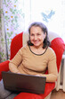 © DariaTrofimova - Joyful elderly lady using laptop and smiling while sitting in armchair