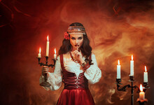 Fantasy Gypsy Woman In Red Vintage Dress. Long Black Hair, Gold Jewelry. Girl Witch Fortune Teller Holds A Candlestick With Burning Candles. Room Is On Fire, Smoke. Spiritual Seance, Summons Spirits.