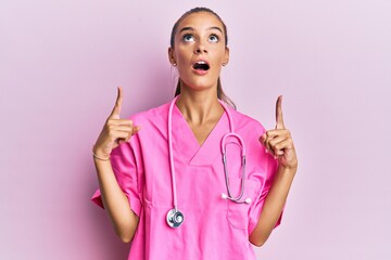 Wall Mural - Young hispanic woman wearing doctor uniform and stethoscope amazed and surprised looking up and pointing with fingers and raised arms.