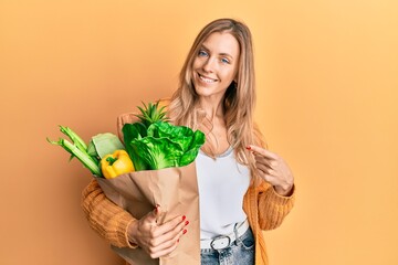 Sticker - Beautiful caucasian woman holding paper bag with bread and groceries smiling happy pointing with hand and finger